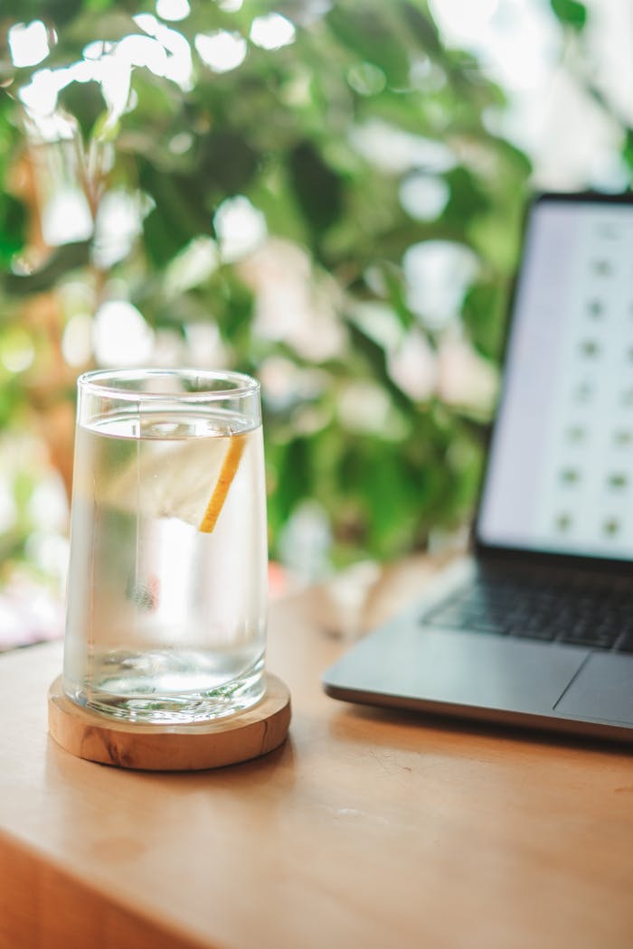 A glass of lemon water beside a laptop on a wooden table, creating a refreshing workspace atmosphere.