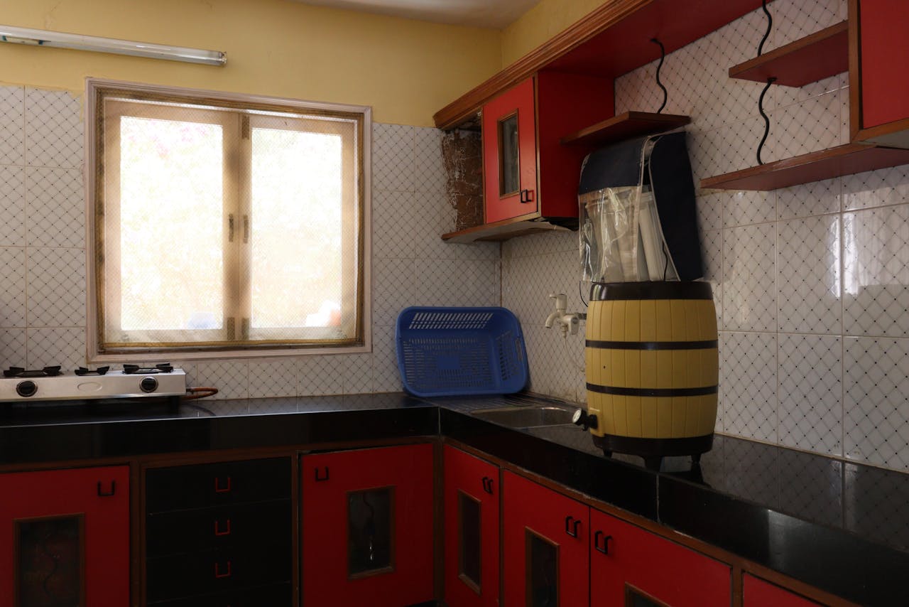 Bright Indian kitchen interior with red cabinets, gas stove, and window light.